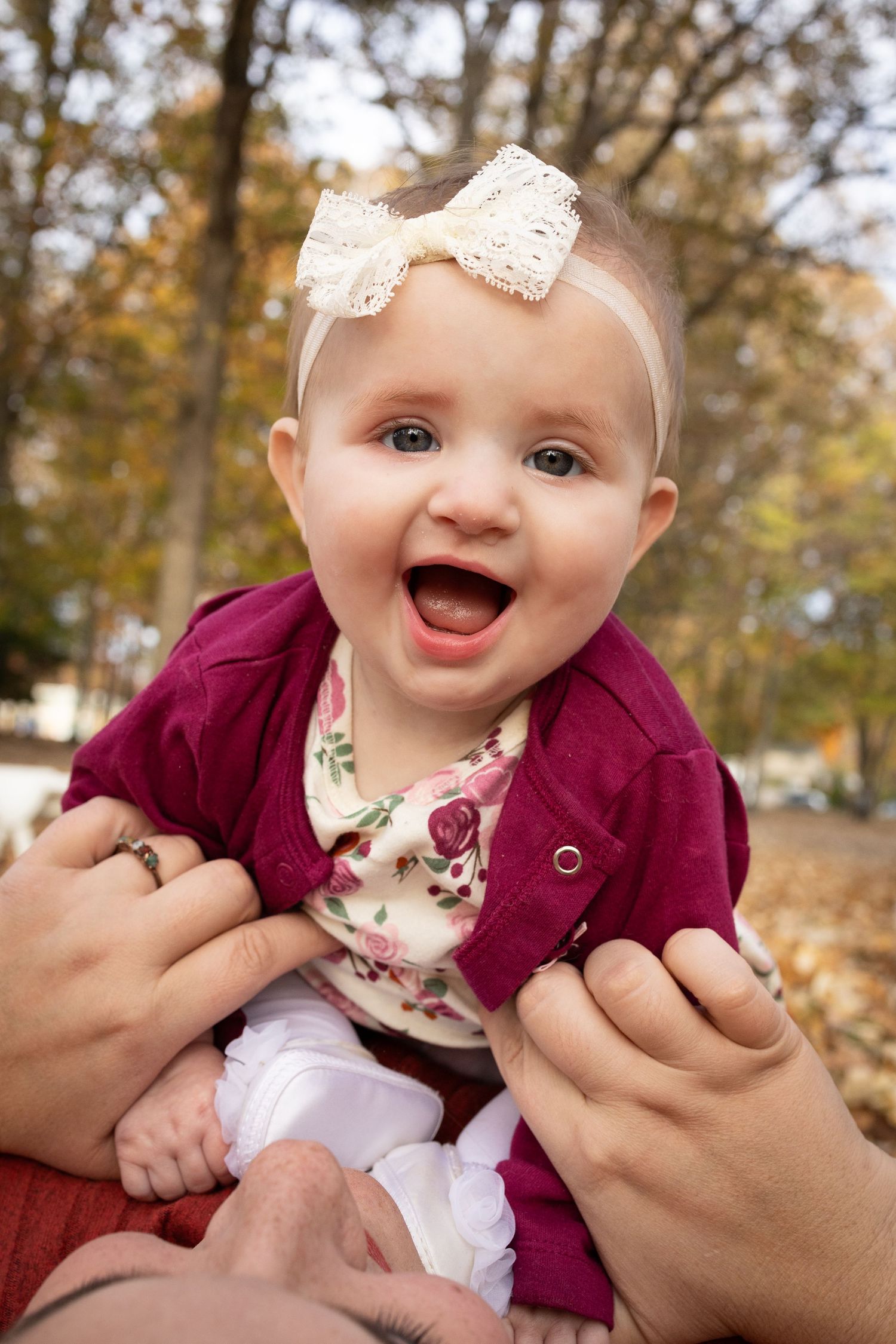 A baby in a burgundy cardigan and floral outfit smiles while being held outdoors in autumn.