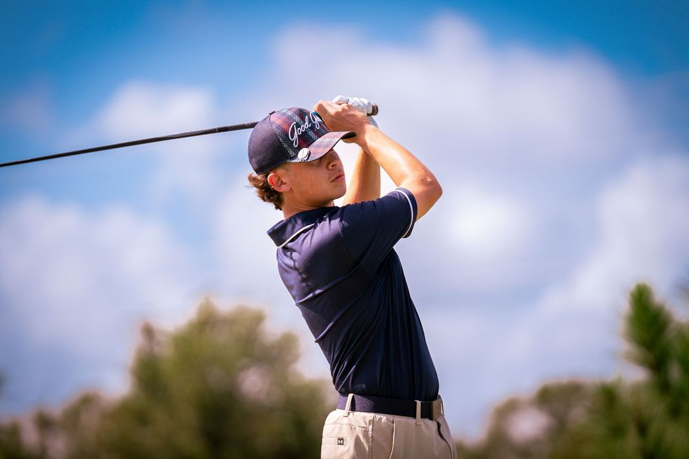 Professional golfer swings club during tournament against blue sky and clouds background.