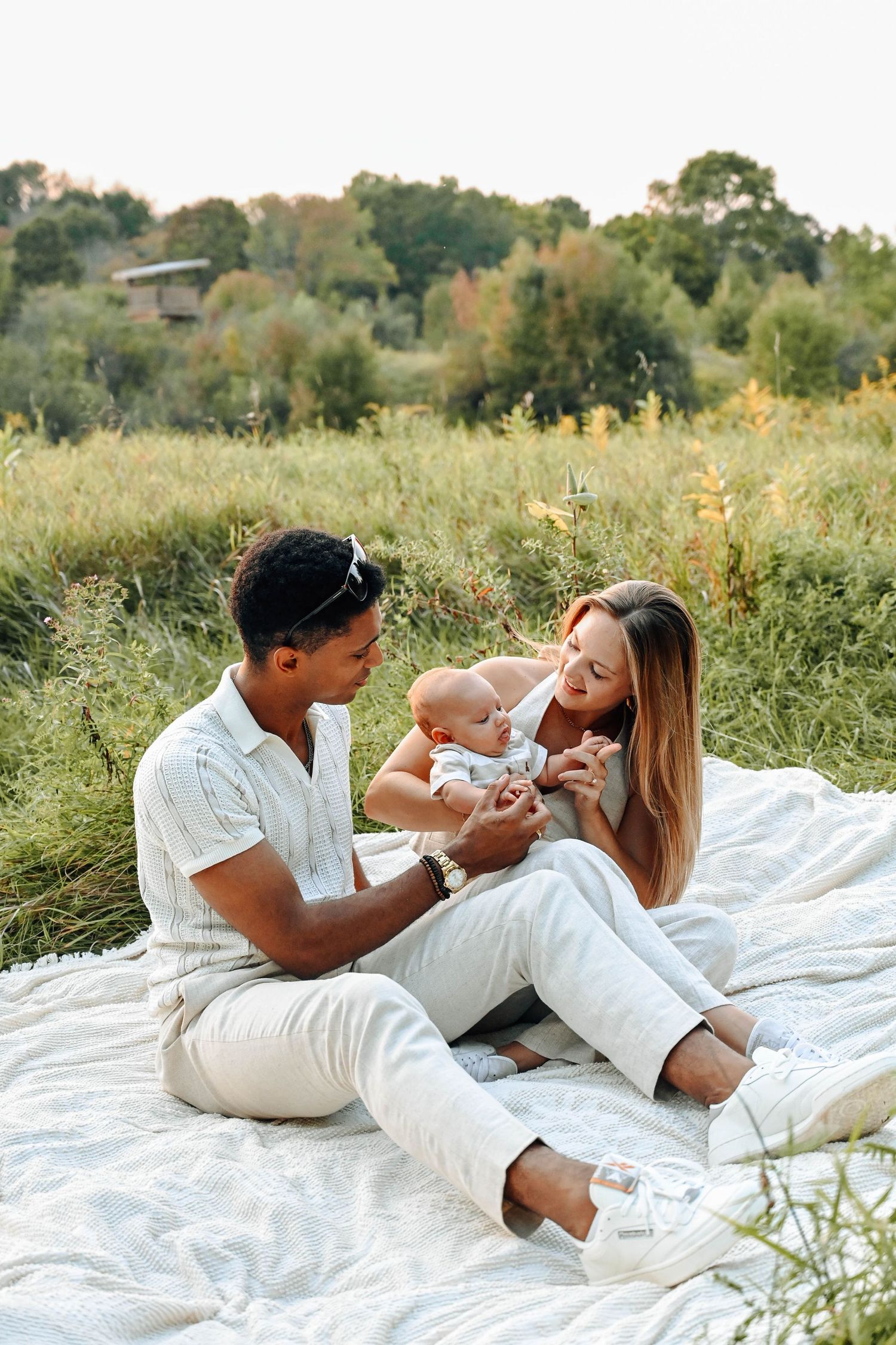 A family picnic scene with parents and baby on white blanket in a sunny meadow.