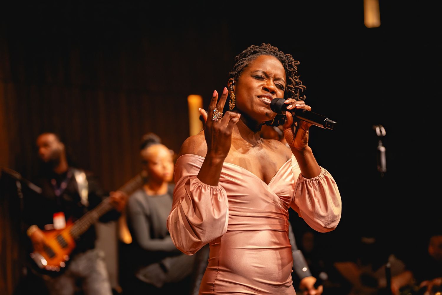 A singer in a pink dress performs passionately on stage with a band visible in the background.