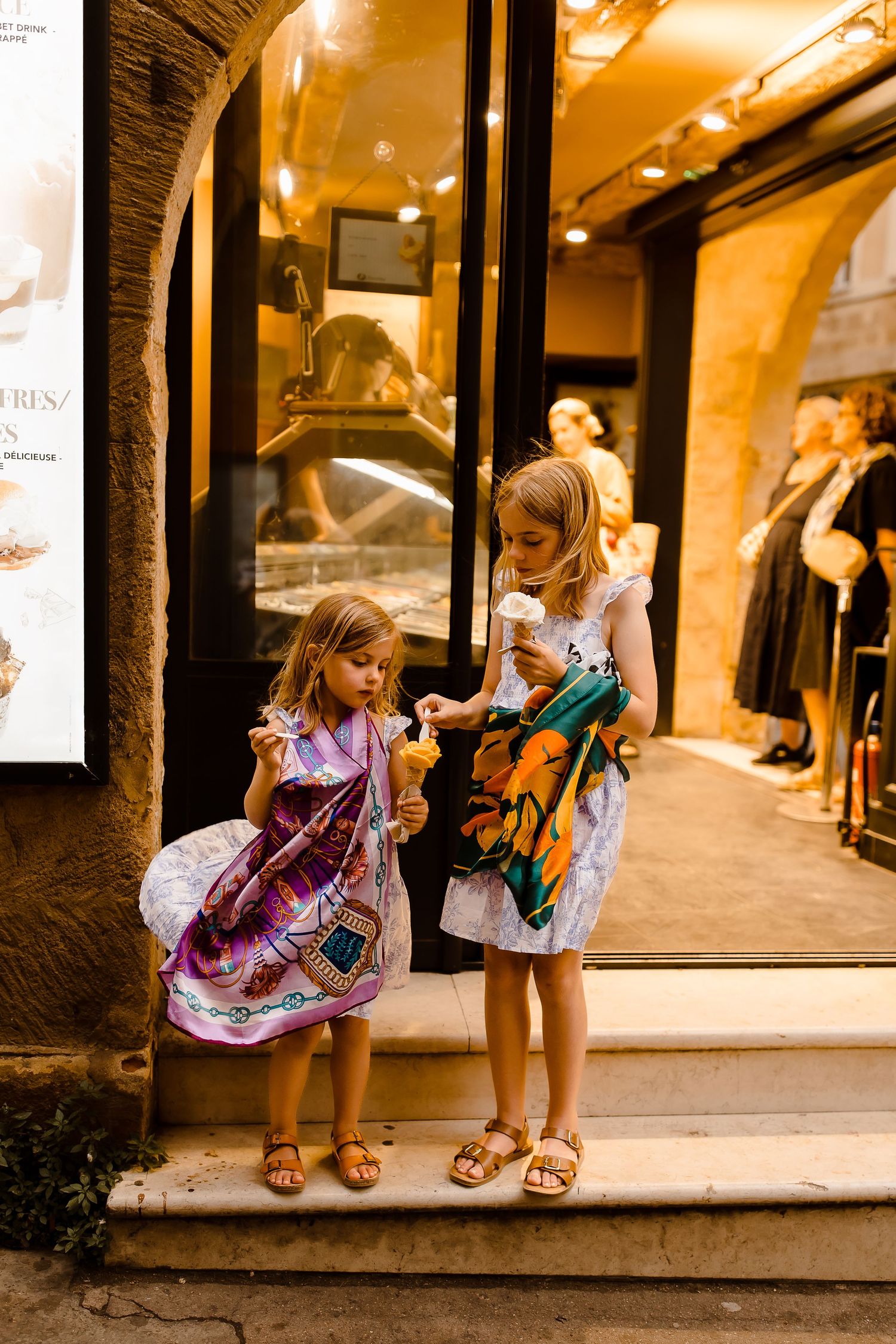 Two children candidly eating ice-cream in front of a warmly lit gelato shop