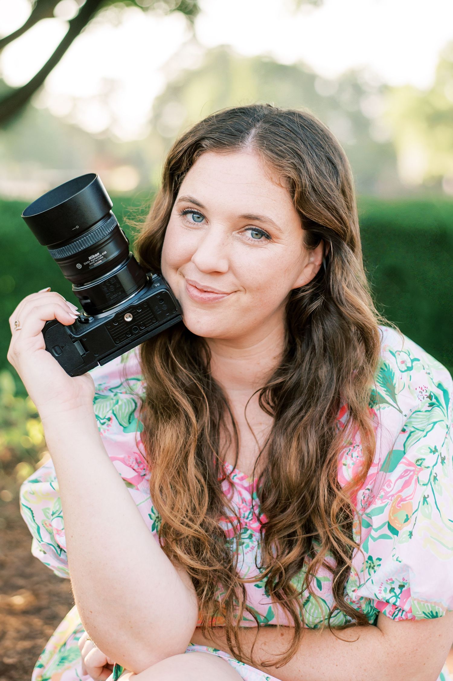 A photographer in a floral dress holds a professional camera while sitting outdoors in natural lighting.