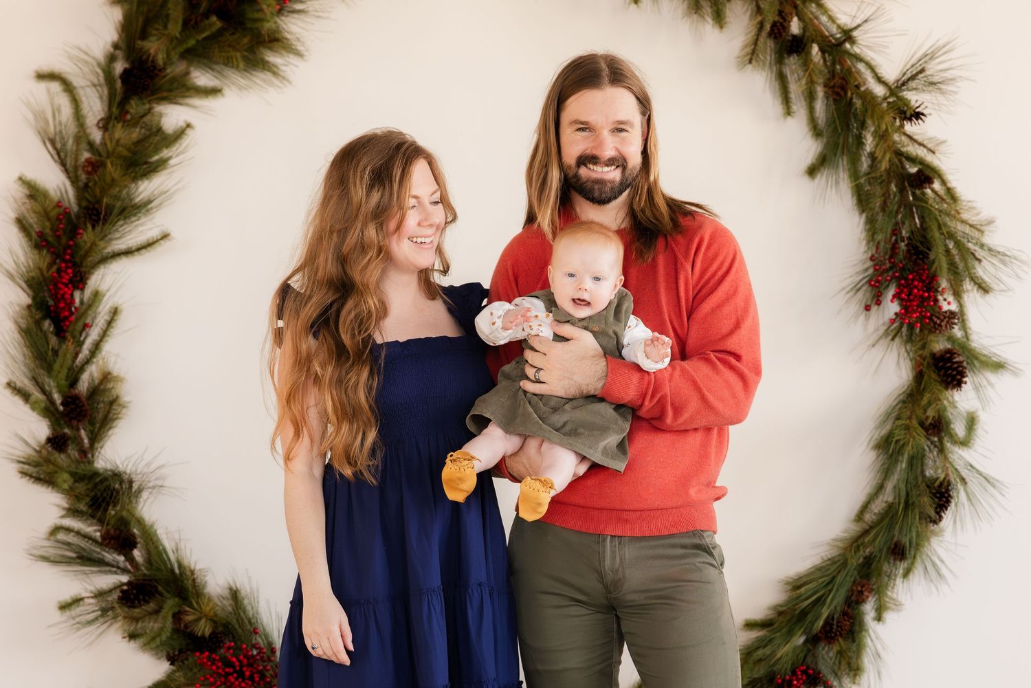Family poses together inside large evergreen wreath Christmas decoration against white wall.