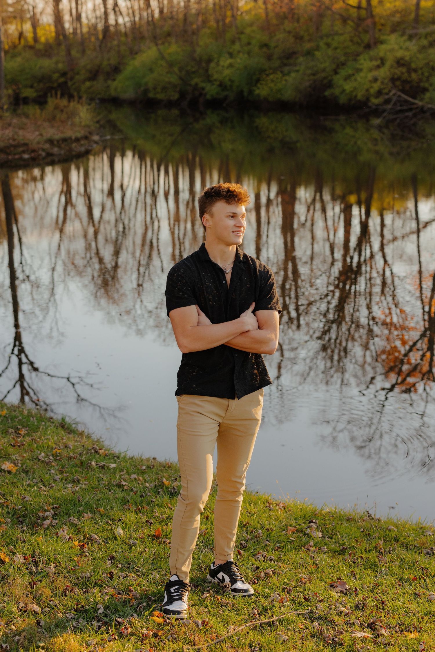 Person in black shirt and khaki pants stands by a tranquil lake with autumn trees reflecting in the water at sunset.