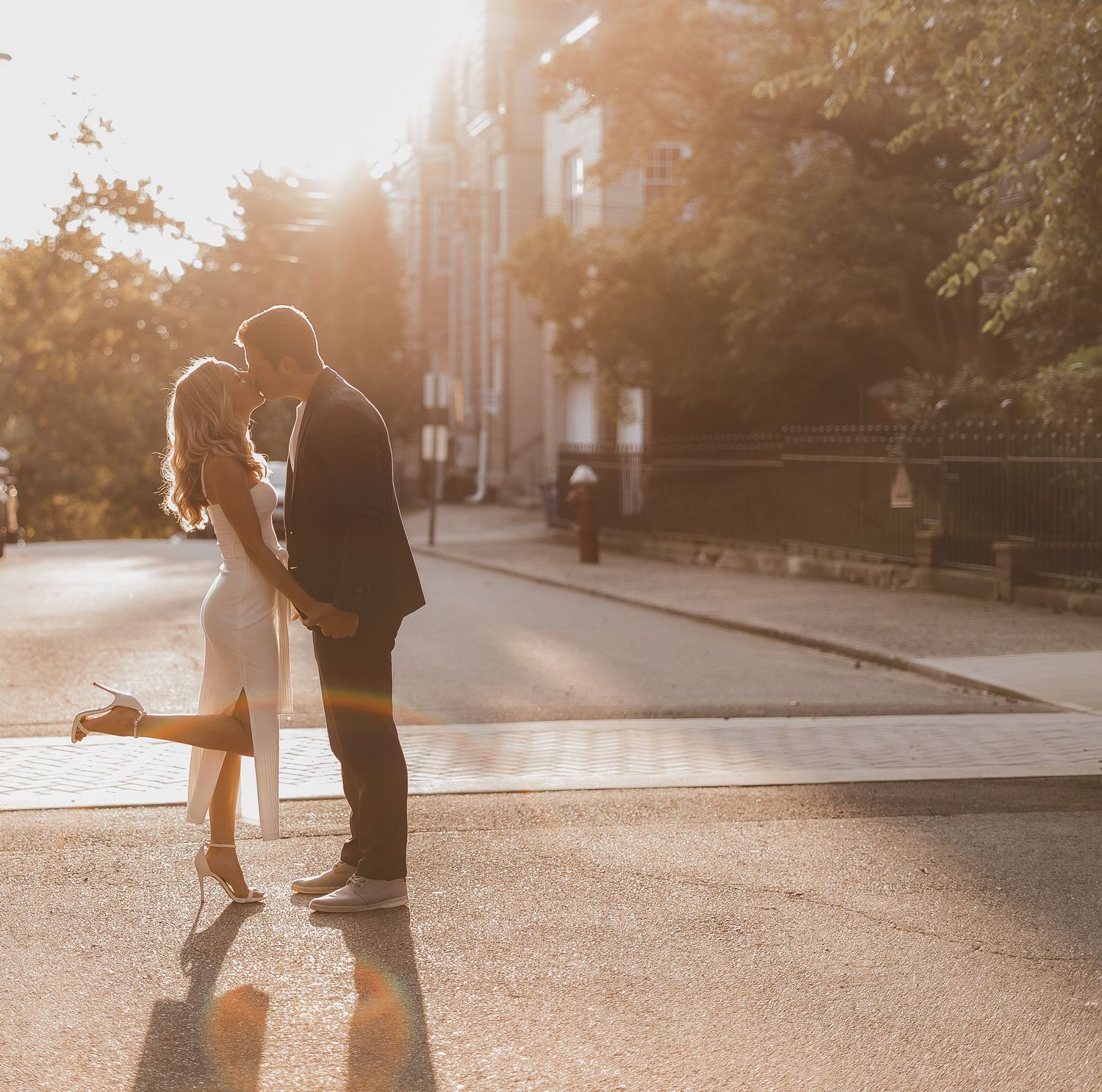 A couple shares an intimate moment on a sunlit city street with golden sunset light in the background.