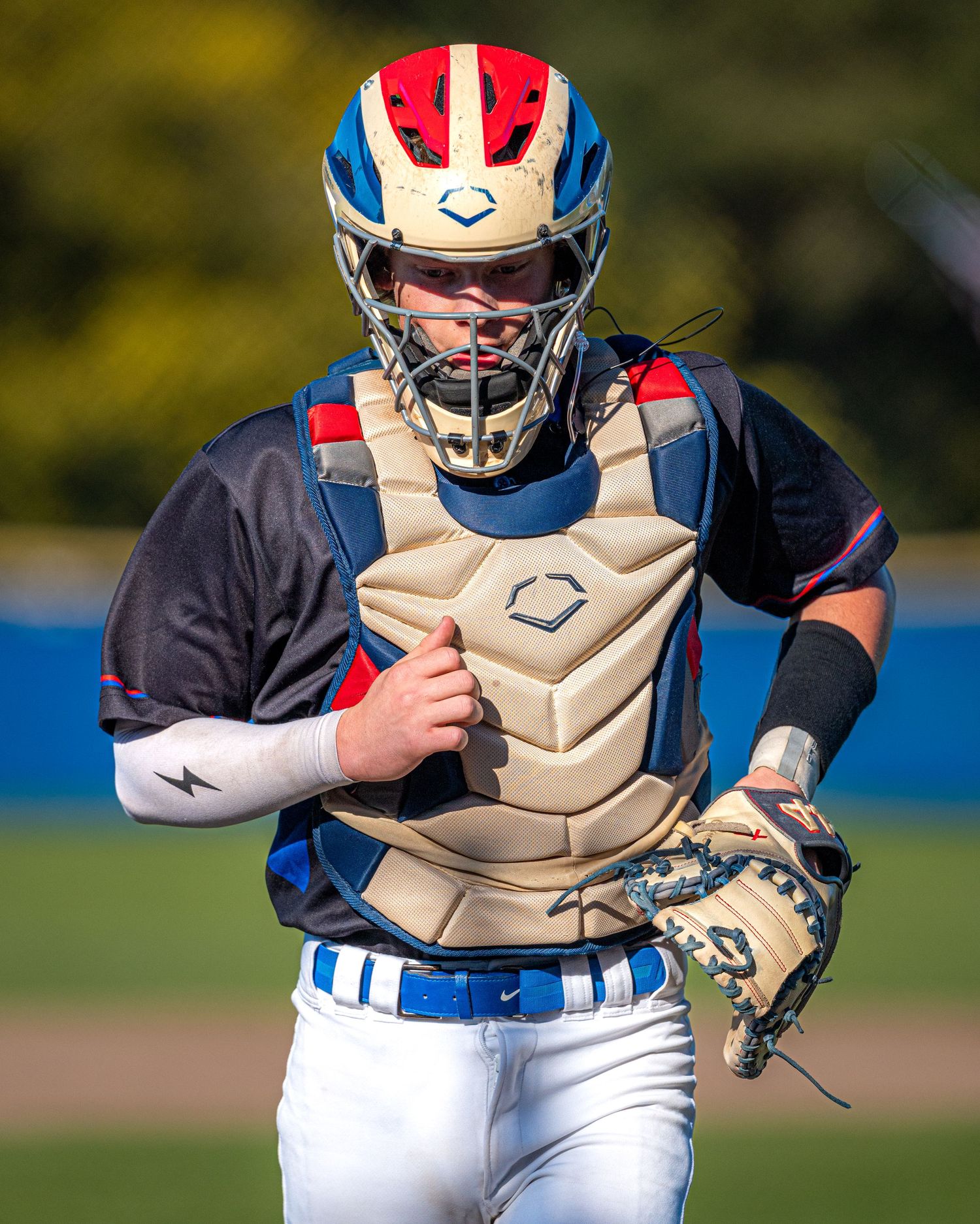Baseball catcher in blue and white uniform with tan protective gear runs on field during sunny day practice session.