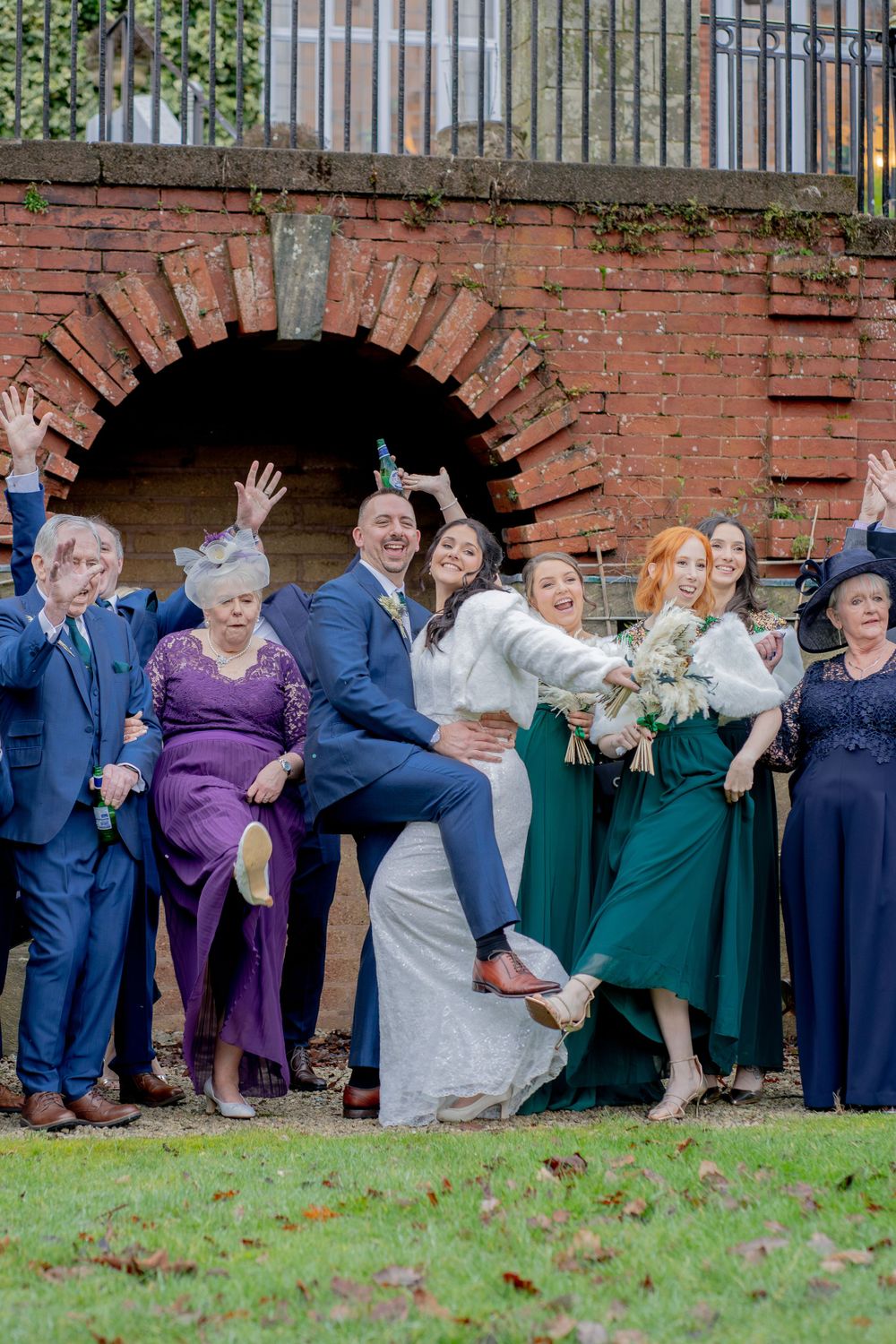 Wedding party poses playfully in front of brick archway with guests wearing purple and emerald green attire.