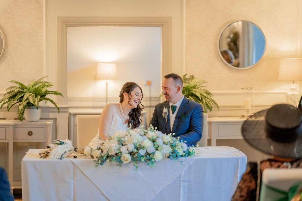 A couple in elegant wedding attire share a moment at a reception table adorned with white and blue floral arrangements.