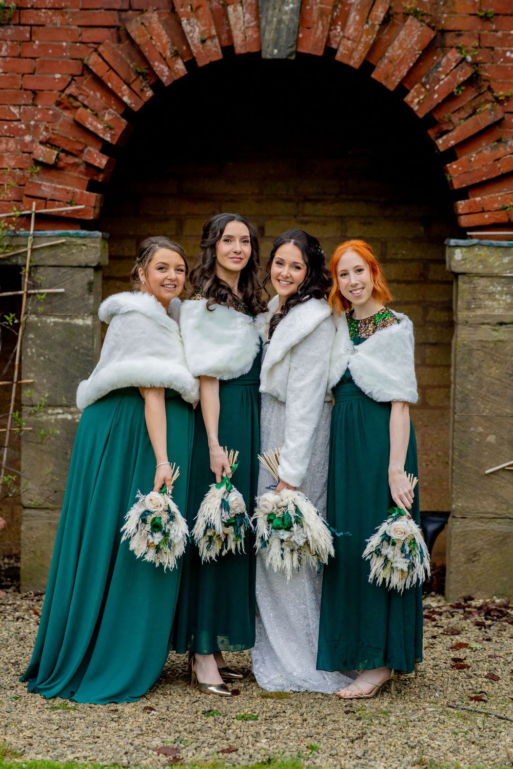 Wedding party in emerald green dresses and white fur stoles hold matching bouquets while posing under brick archway.