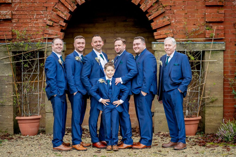 Wedding party in matching navy blue suits poses against a brick archway backdrop.