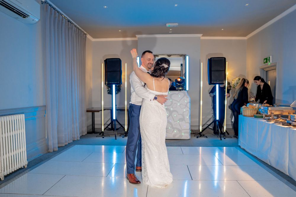 A couple shares their first dance at their wedding reception with DJ speakers and a buffet table visible in the background.