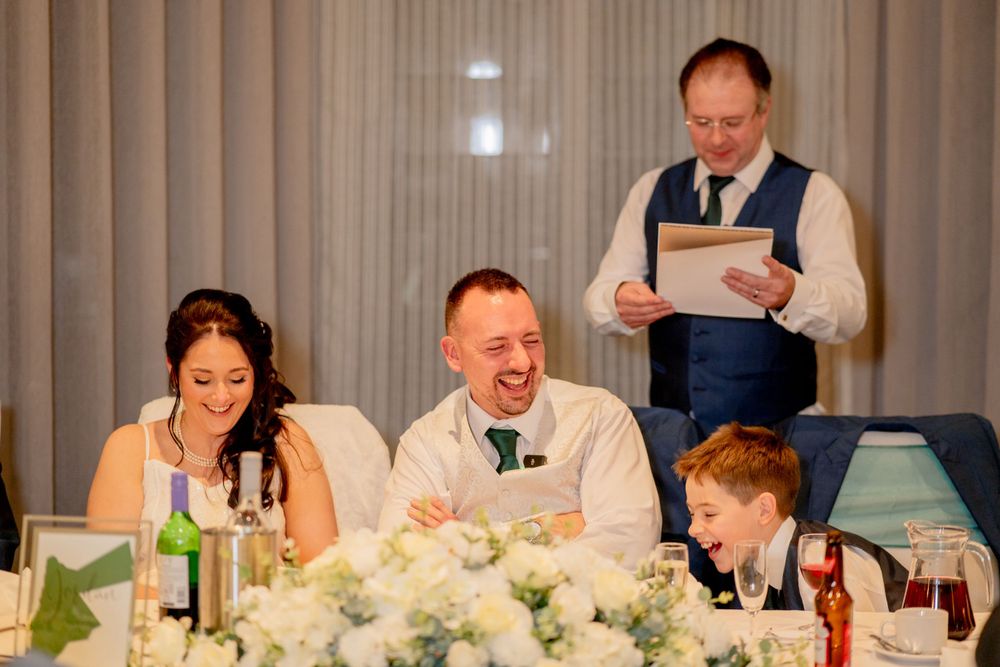Wedding reception scene with guests laughing at dinner table decorated with floral centerpieces.