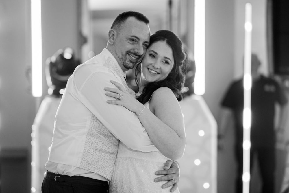 A black and white romantic photograph of a couple embracing and smiling during a slow dance at a wedding celebration.