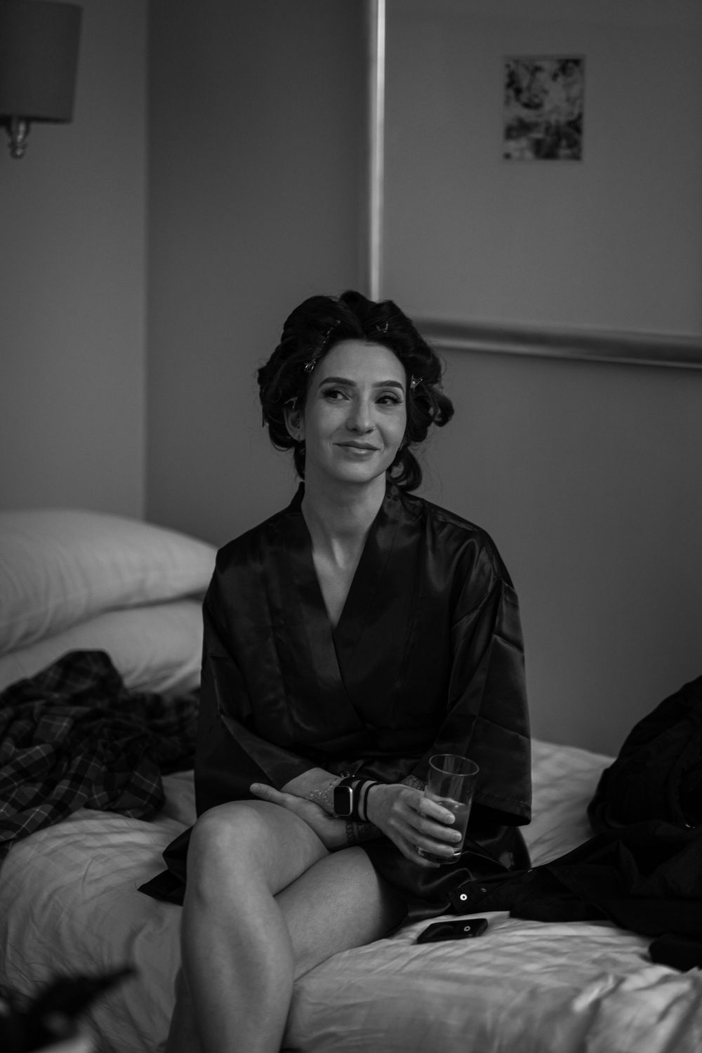 A woman in a black silk robe sitting on a bed during wedding preparations.