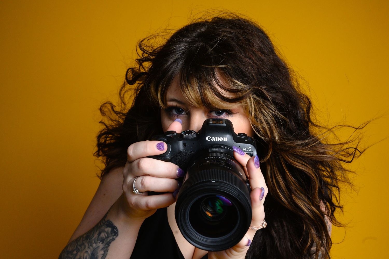 Female photographer aiming a Canon camera directly at viewer against a bold yellow background.