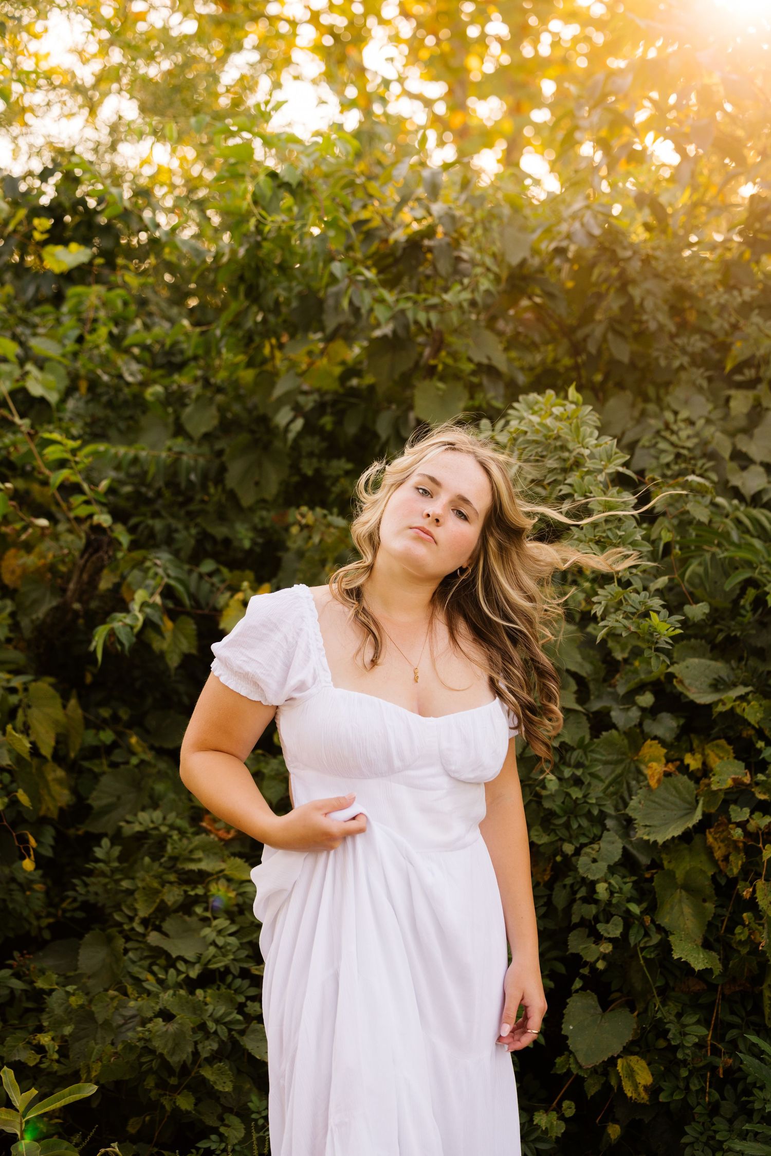 A woman in a flowing white dress poses outdoors at sunset with foliage in the background.