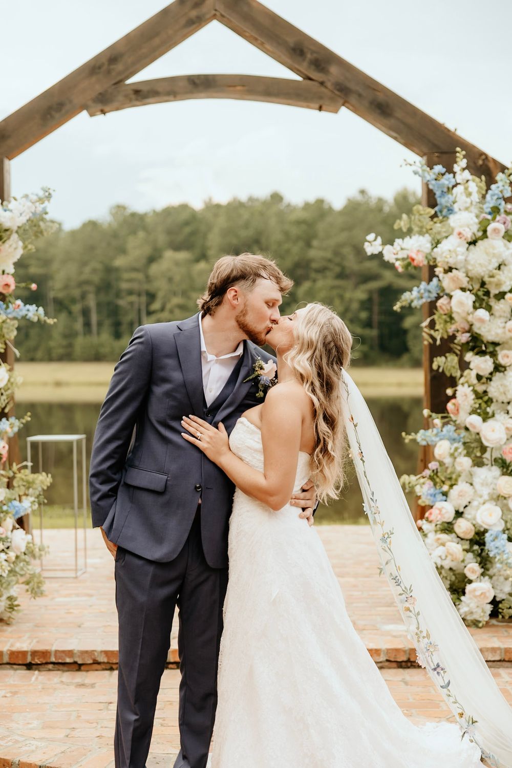 Newlyweds share a romantic kiss under a rustic wooden arch decorated with white floral arrangements by a lakeside.