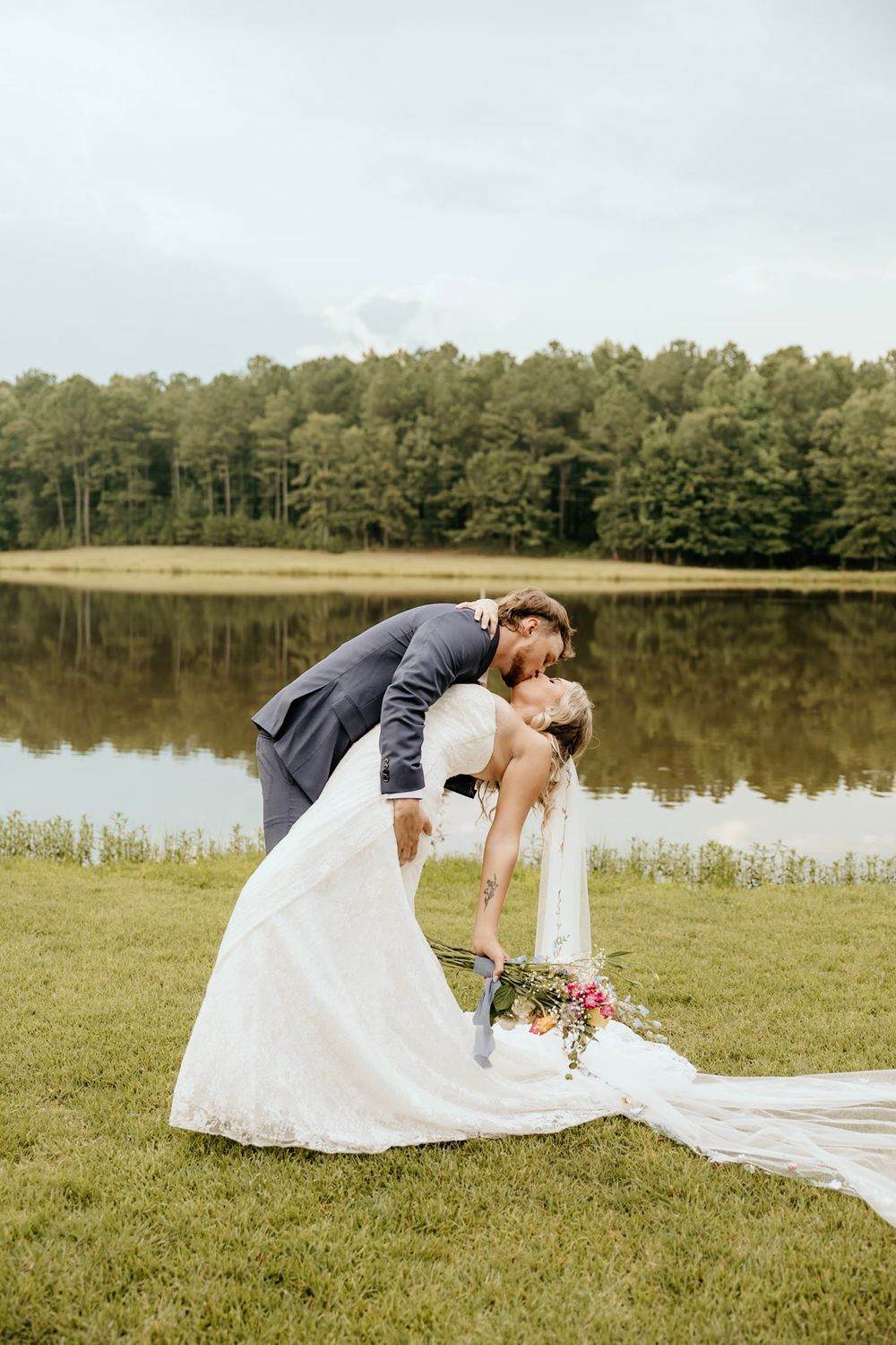 Romantic wedding photo showing elegant couple in formal attire embracing by serene lake with pine forest backdrop.