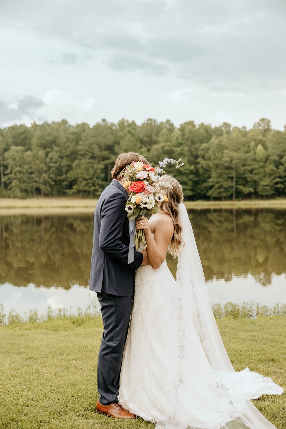 A romantic wedding couple shares a kiss by a serene lake with trees in the background.