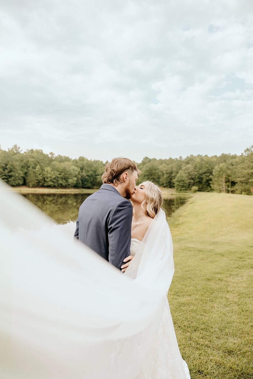 A romantic wedding couple embraces in a beautiful outdoor setting with flowing white veil and green landscape.