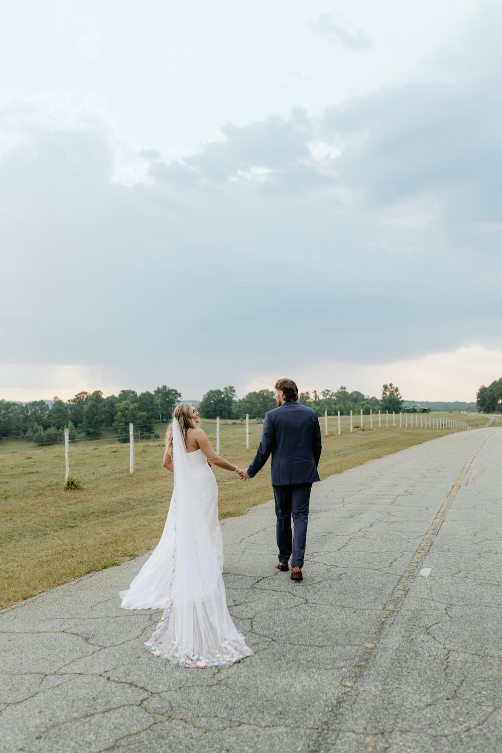 A couple in wedding attire holding hands while walking down a rural concrete path surrounded by fence posts and fields.