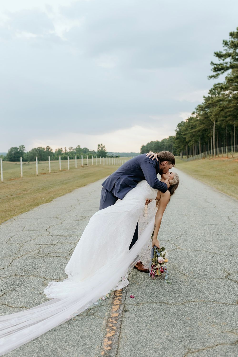 A romantic dip kiss between newlyweds on a long path lined with trees and fencing.