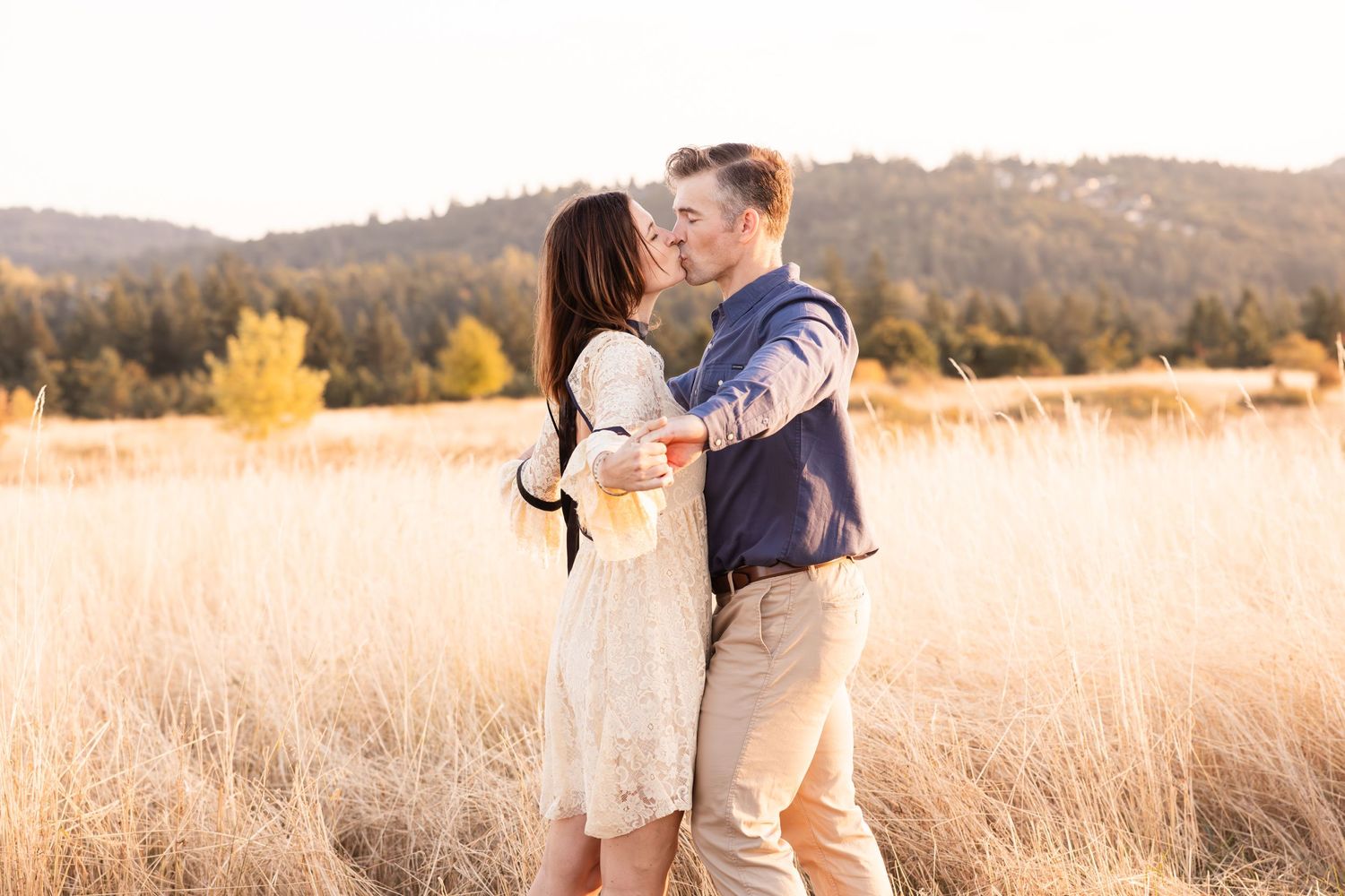 Couple embracing in a field with mountains and autumn trees in the background.