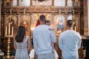 Three people facing ornate Orthodox iconostasis with religious artwork and gold details.