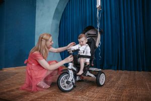 A child sits on a tricycle while someone in coral dress assists.
