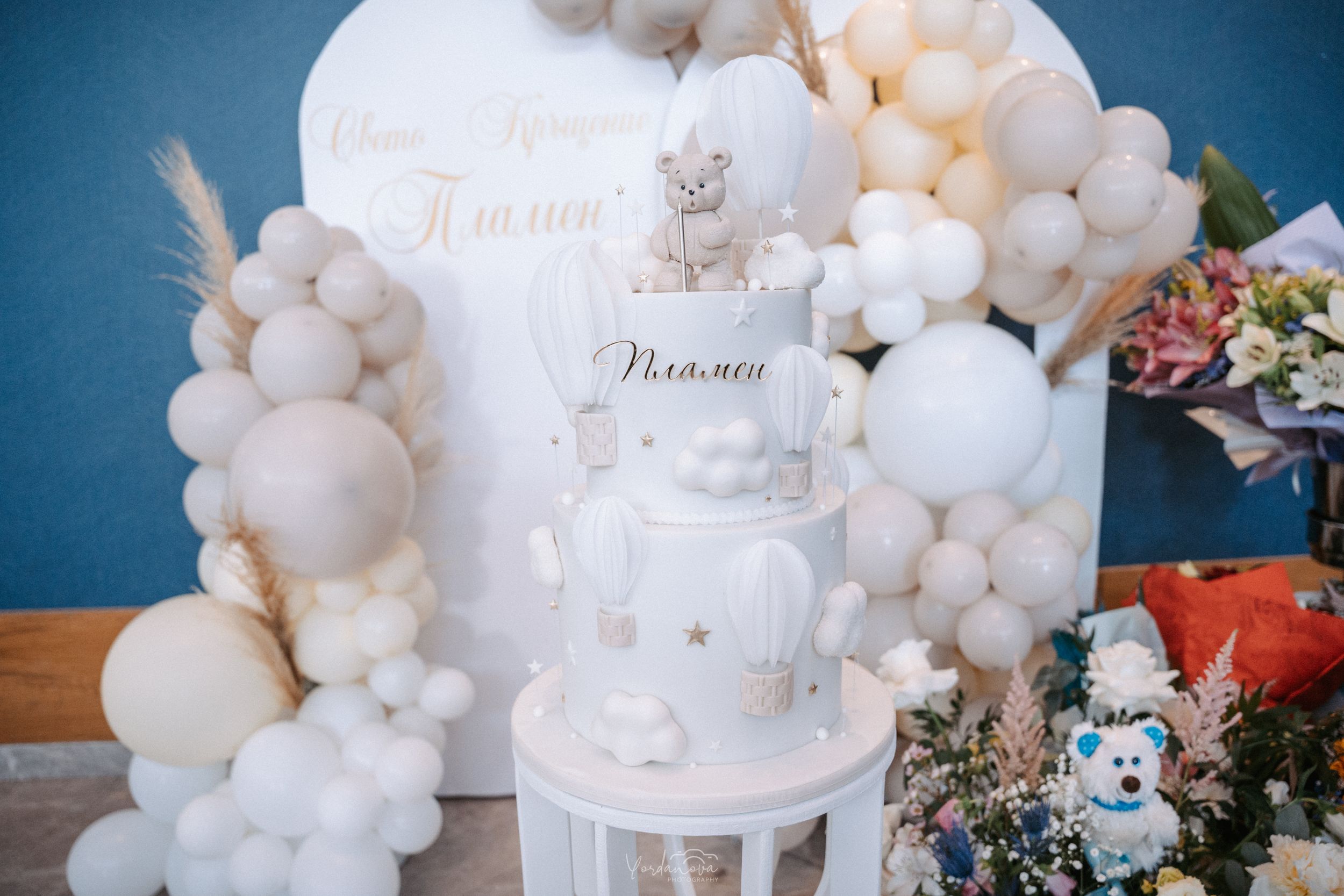 White tiered celebration cake with balloon decorations and floral accents.