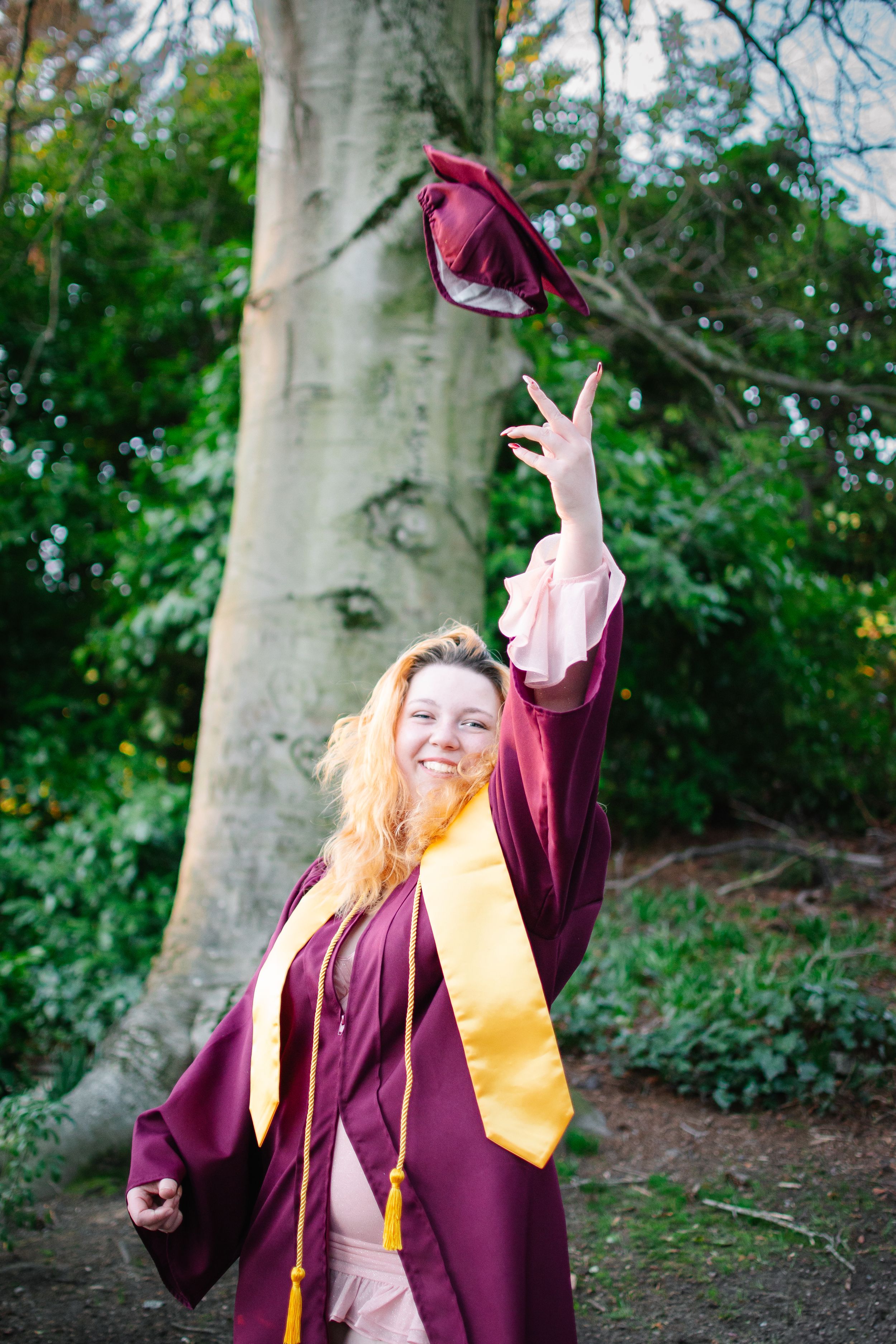High school senior in maroon and gold cap and gown holding graduation cap up with a smile during golden hour at Kerry Park in Seattle.
