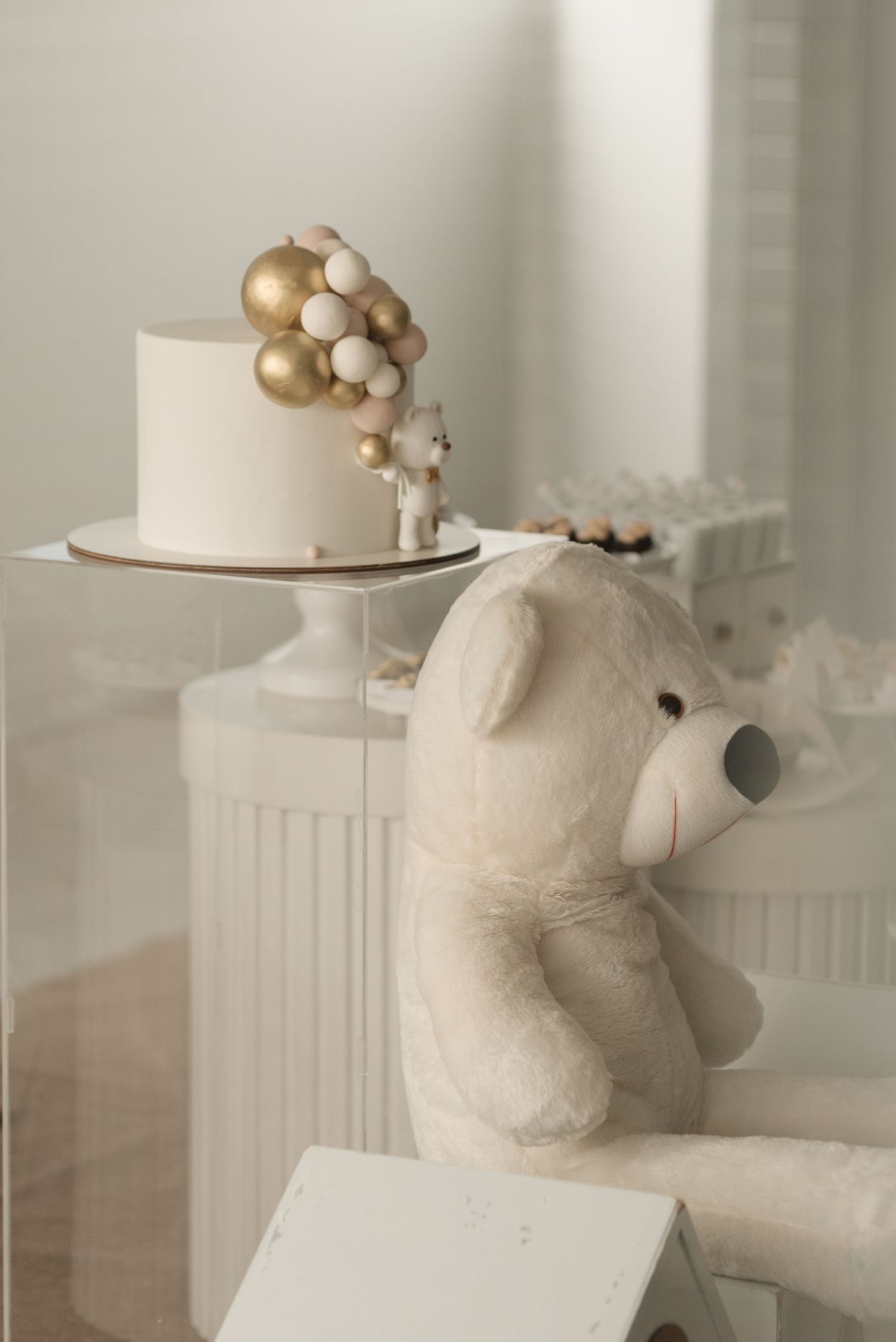 White cake with gold balloon decorations and plush teddy bear on elegant display.