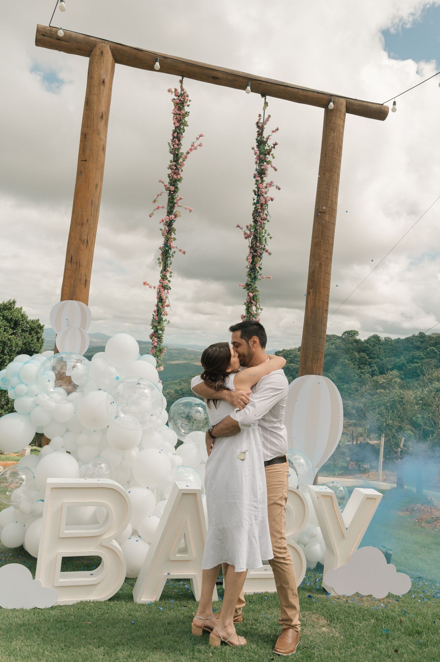Romantic couple kissing near wooden arch with white balloons and BABY letters outdoors.