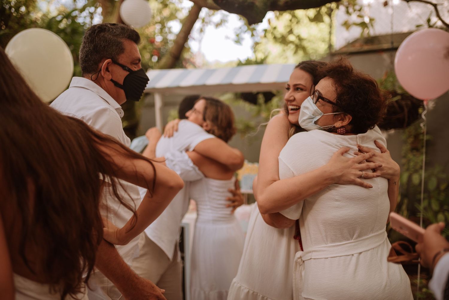 A celebratory gathering outdoors with white and pink balloons where guests share warm embraces.