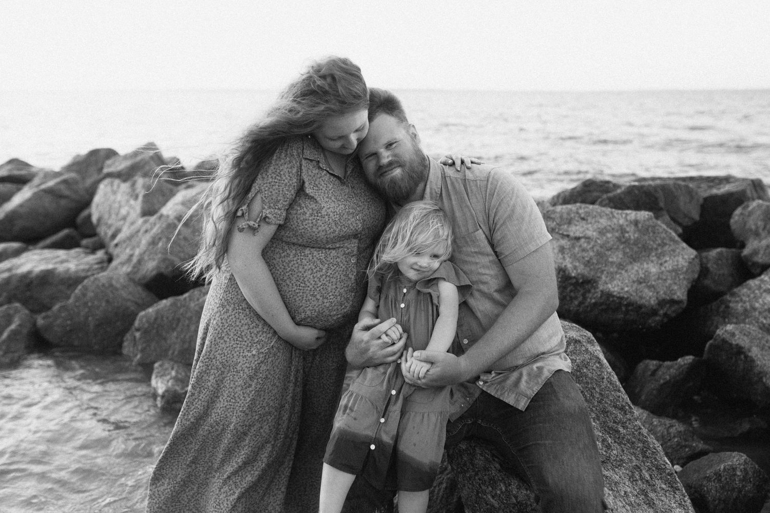 Photo by B.Logsdon of a family sharing a tender moment while sitting on coastal rocks by the HHI ocean at sunset in black and white.