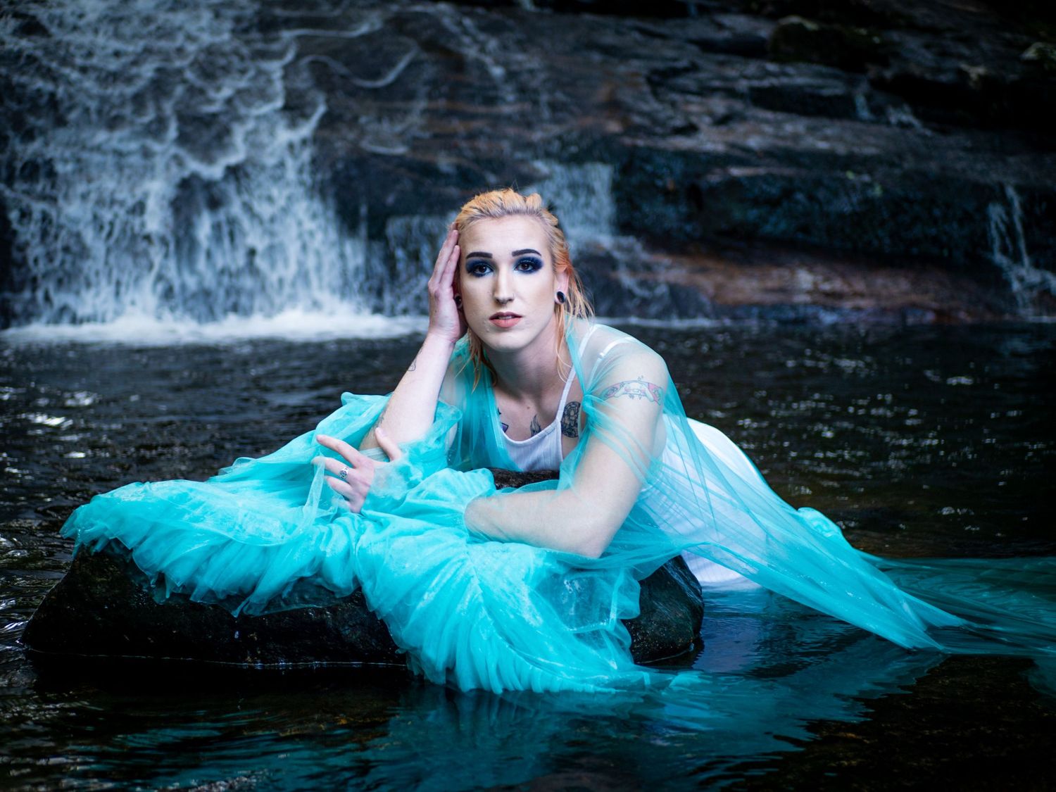 A figure in a turquoise tulle dress poses dramatically in dark water with a waterfall backdrop.