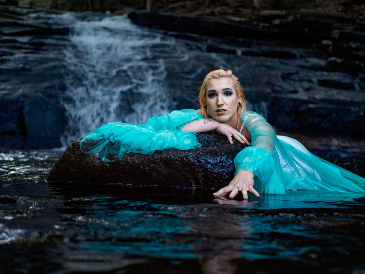 Woman in flowing turquoise dress poses dramatically in dark waters by cascading waterfall.