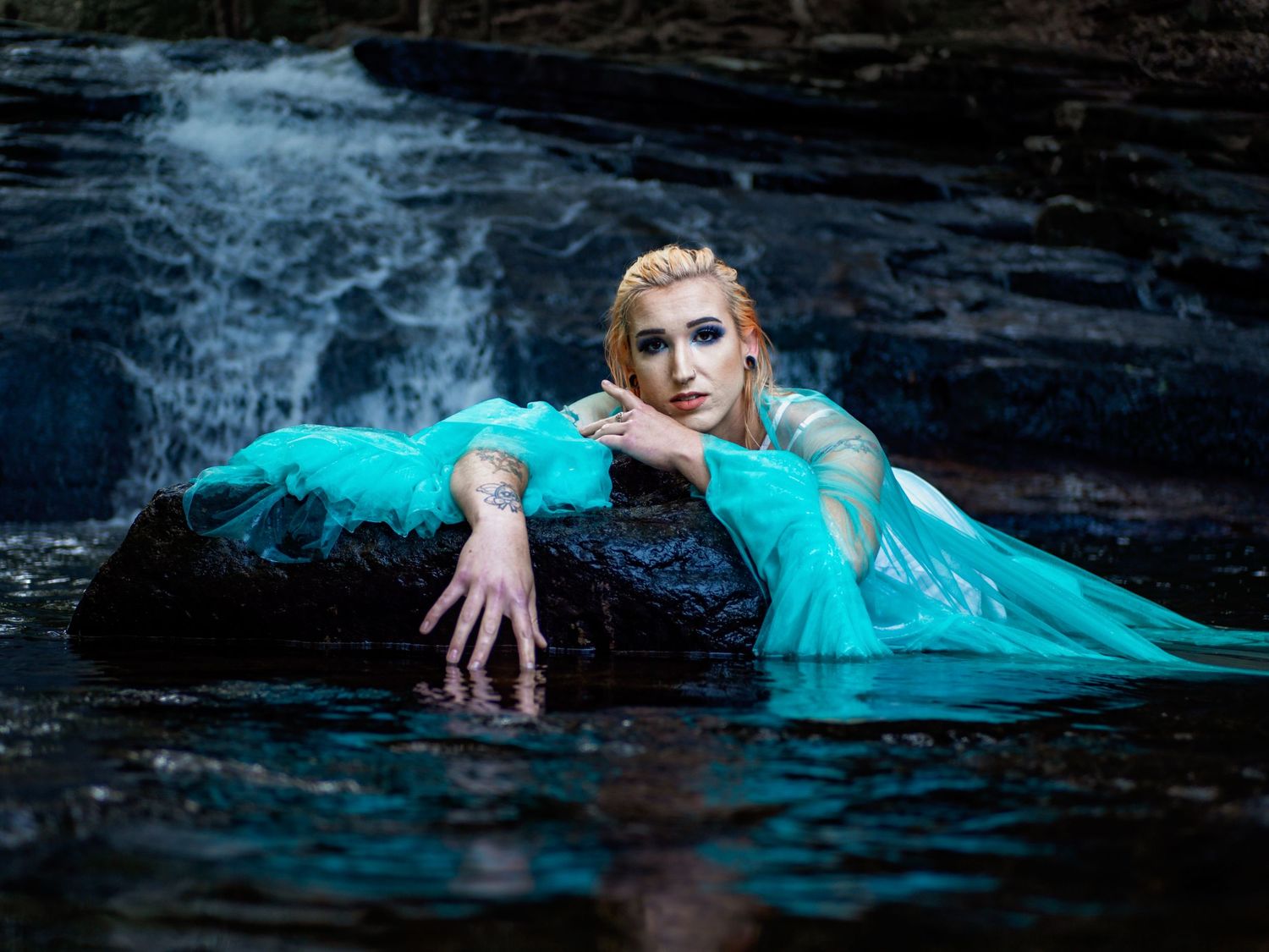 Figure in flowing turquoise dress poses dramatically against dark rocks near rushing water in fantasy-inspired portrait.