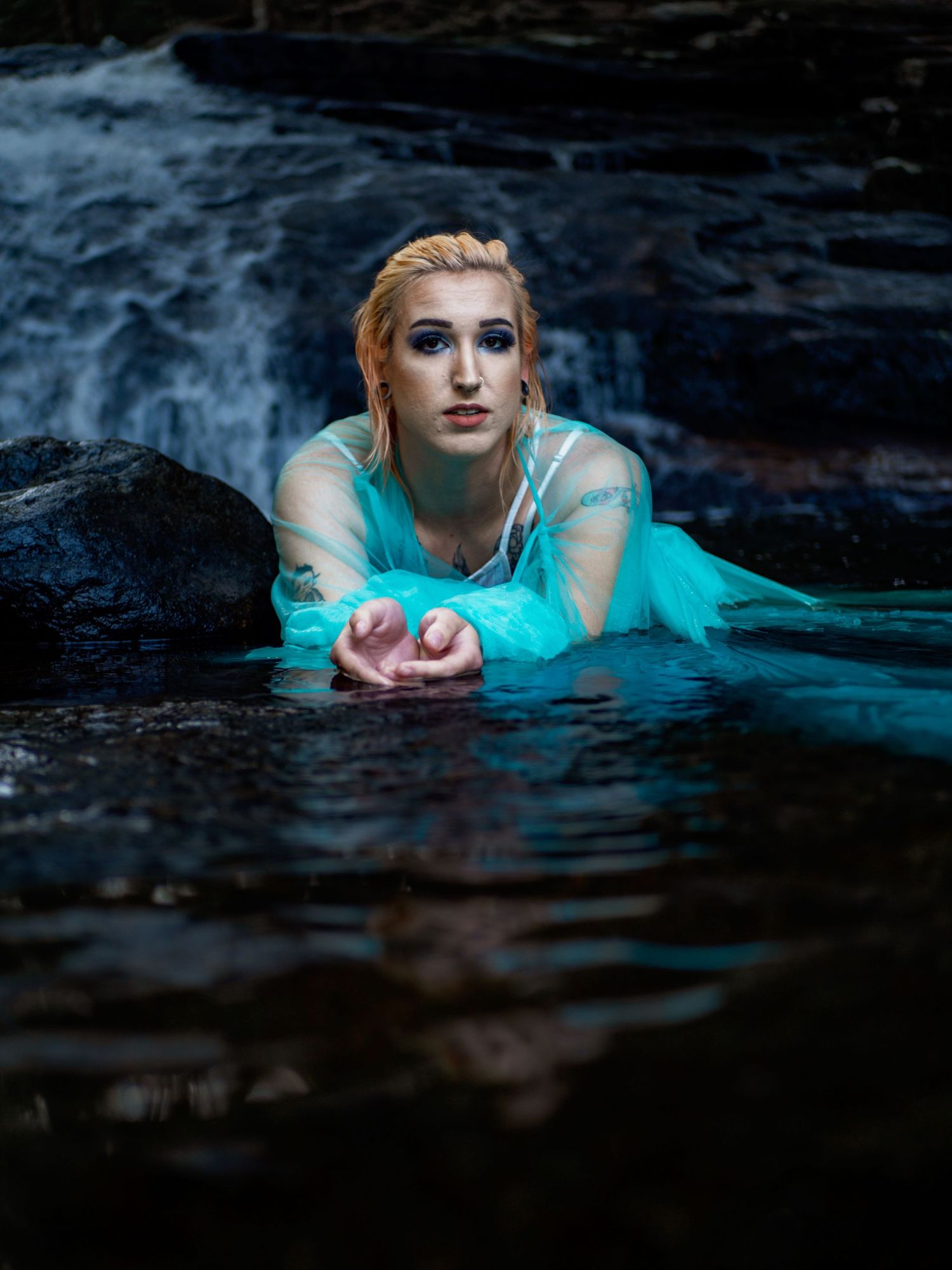 A person in a turquoise dress lying in dark flowing water against a moody background.