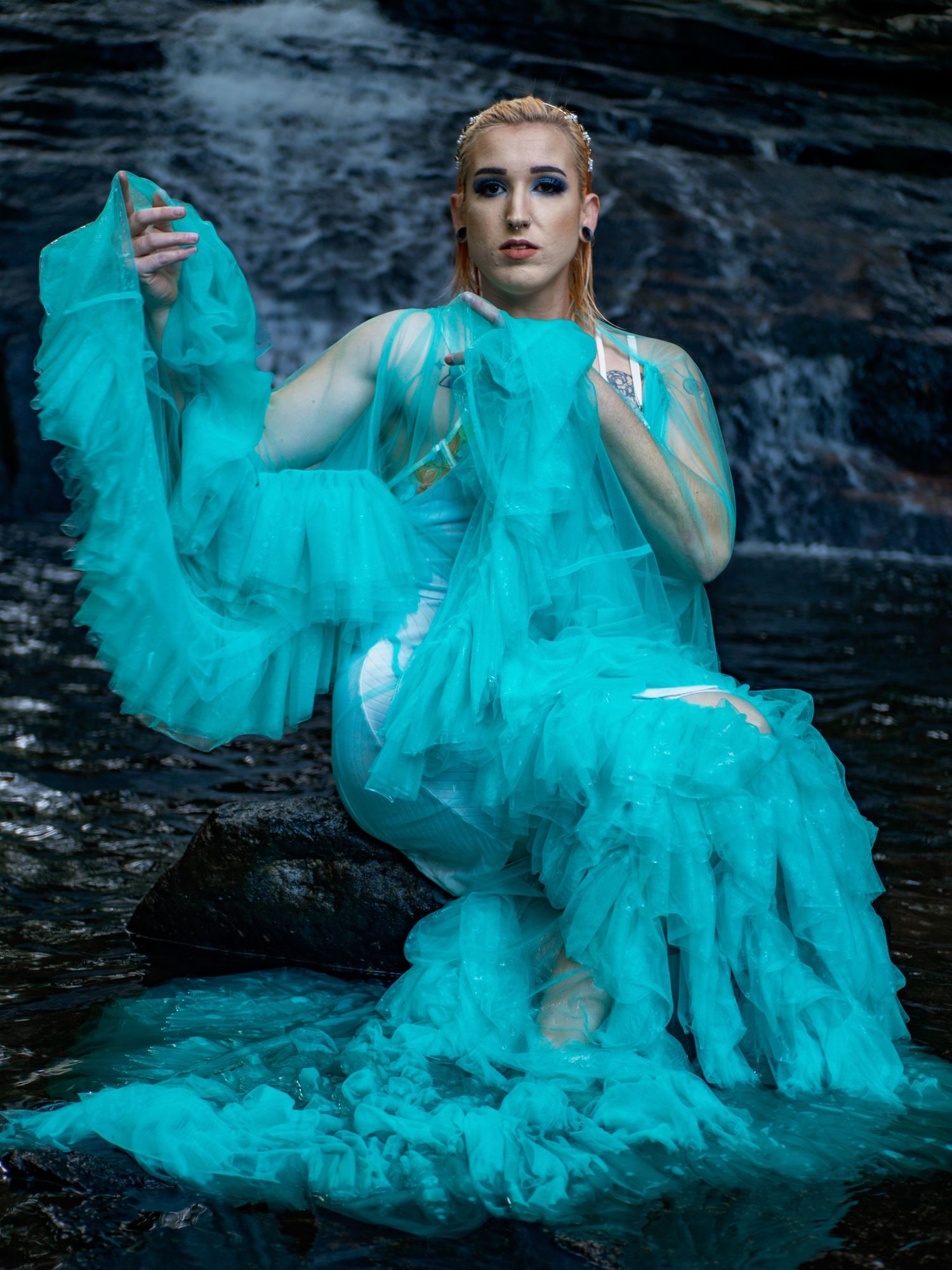 Fashion photography featuring flowing turquoise tulle gown against dramatic waterfall backdrop in moody lighting.