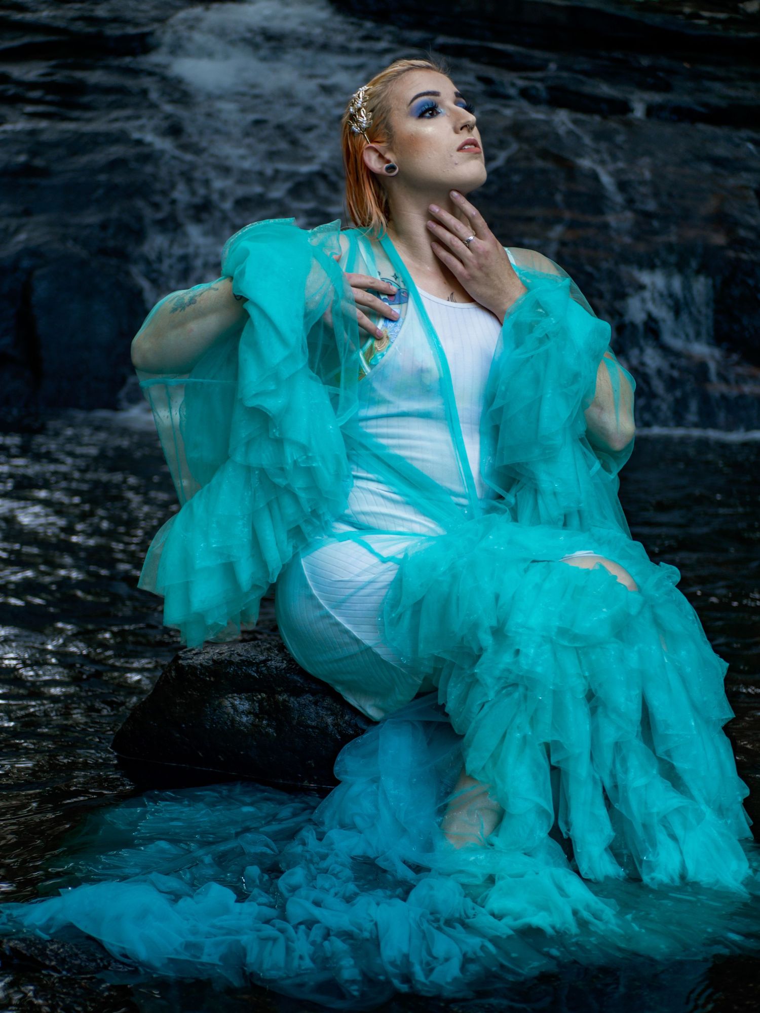 An ethereal portrait of someone wearing a white dress with flowing turquoise fabric posing by a waterfall.