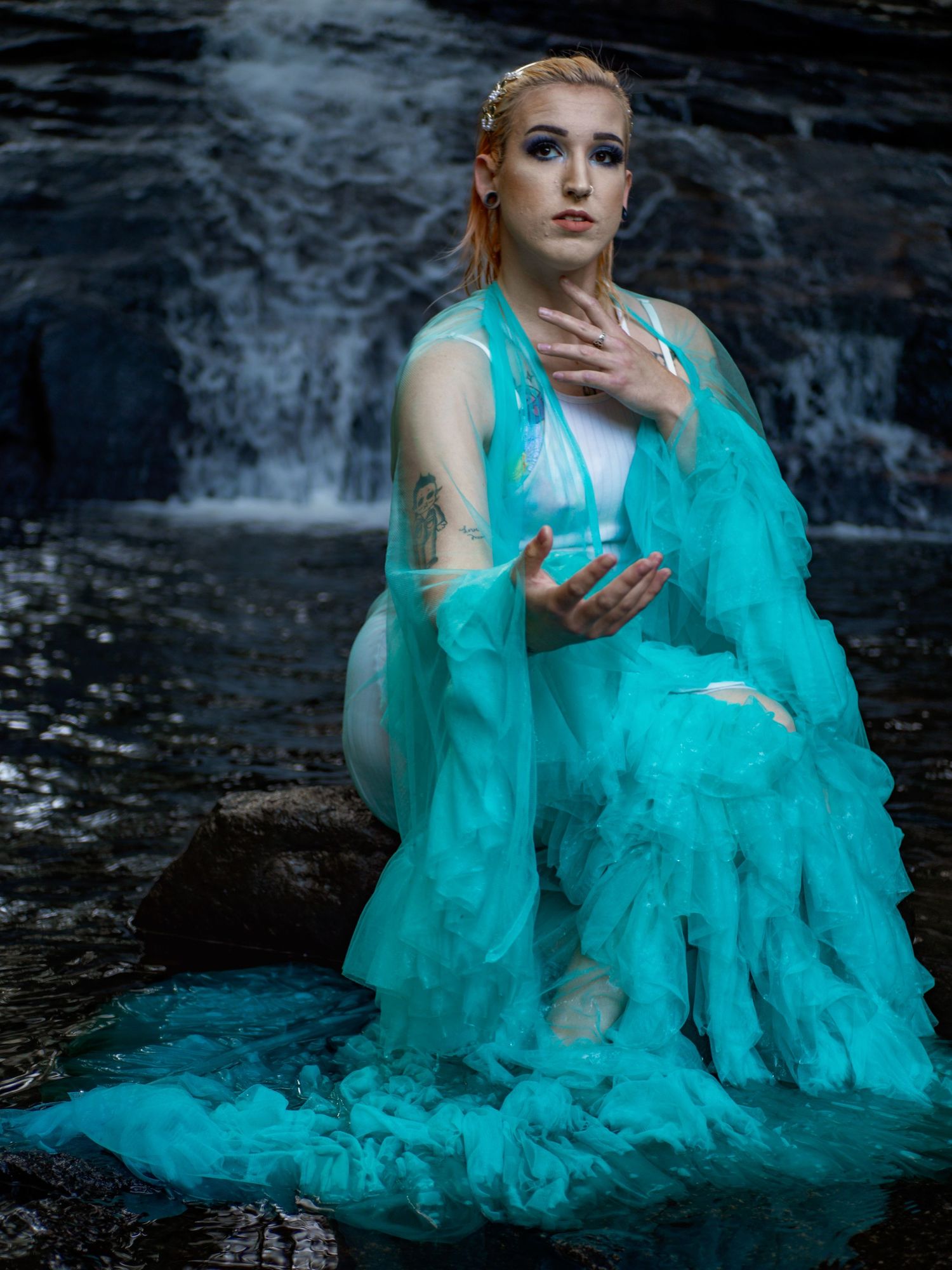 Figure in flowing turquoise dress poses dramatically by waterfall creating ethereal fantasy portrait effect.