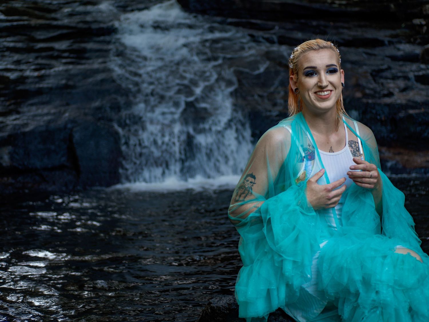 Person in turquoise tulle dress holding a drink while sitting near dark waterfall background.