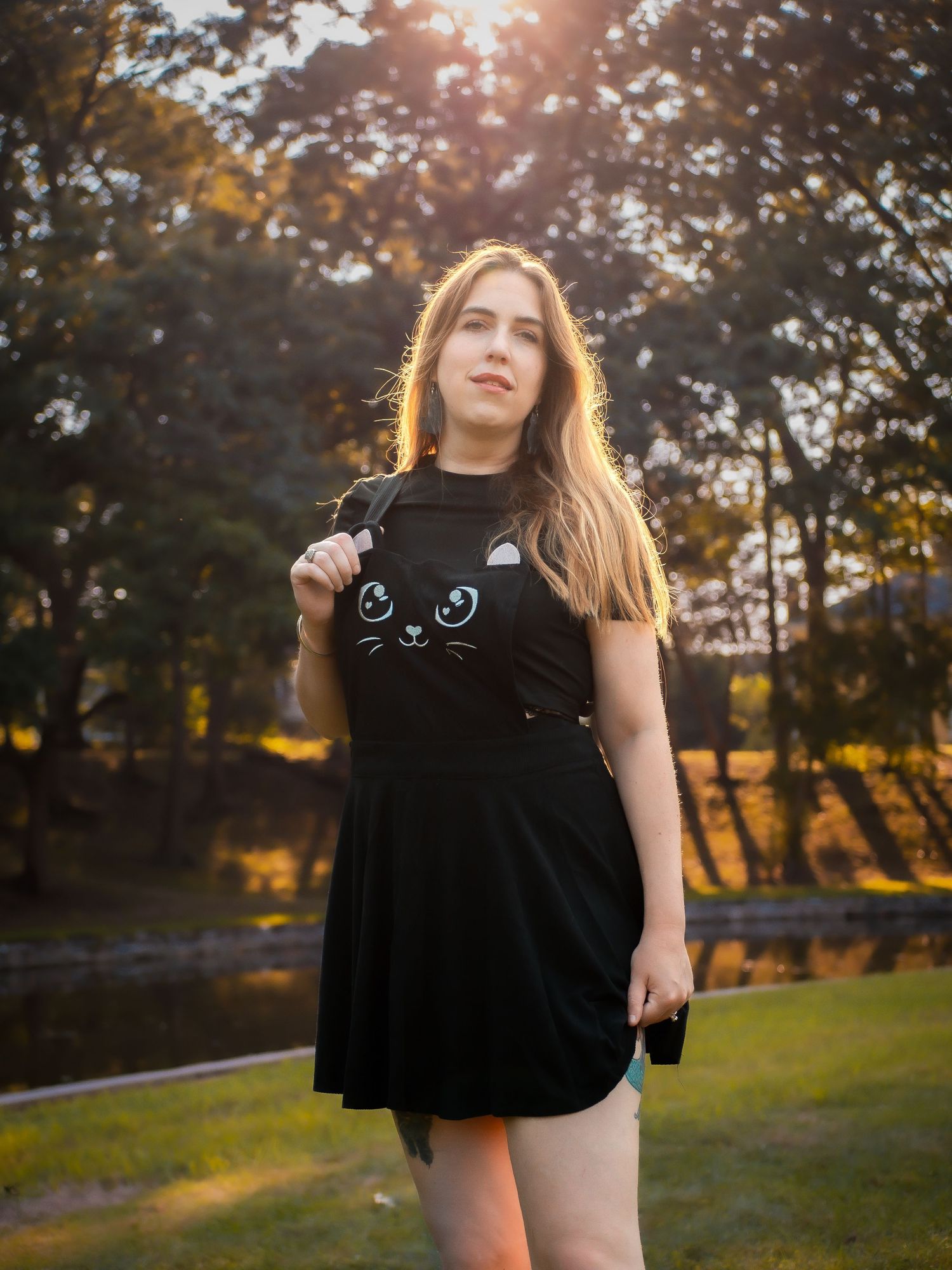 A person in a black cat-themed dress poses outdoors at sunset with golden sunlight filtering through autumn trees.