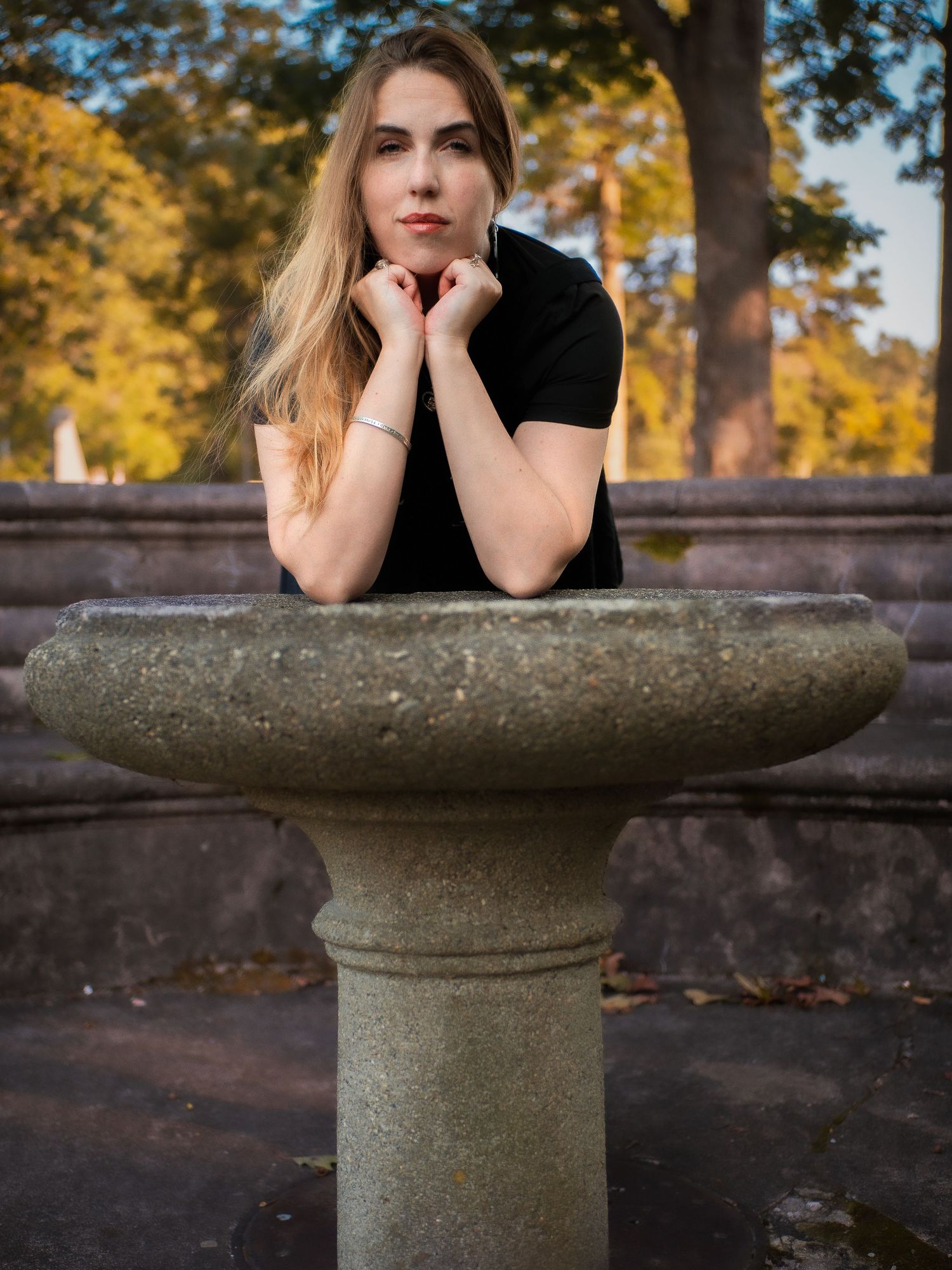 A contemplative pose at a stone birdbath in a park during fall with golden trees in the background.