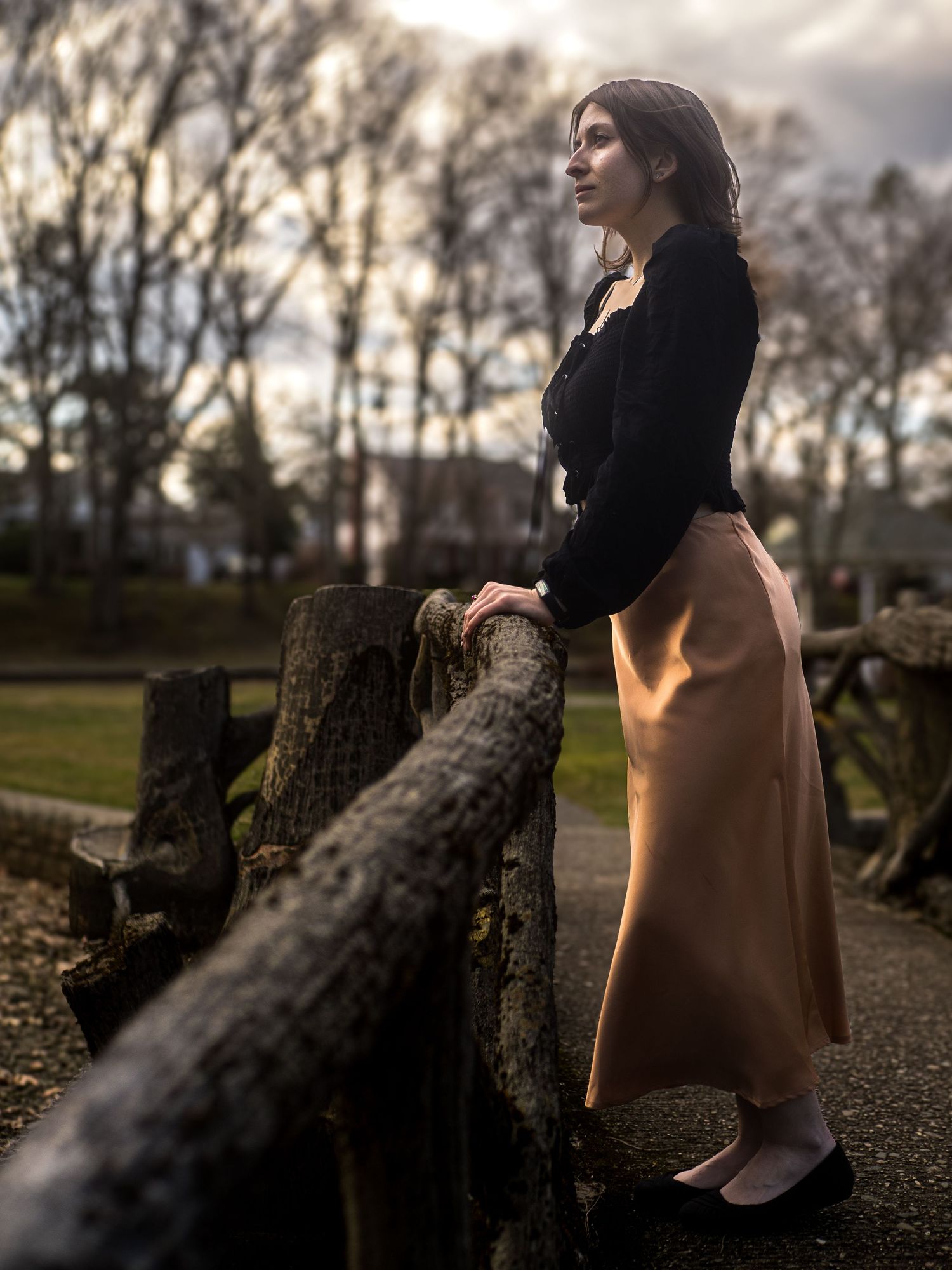 A woman in a black sweater and tan skirt leans against a rustic wooden fence at dusk in a garden setting.