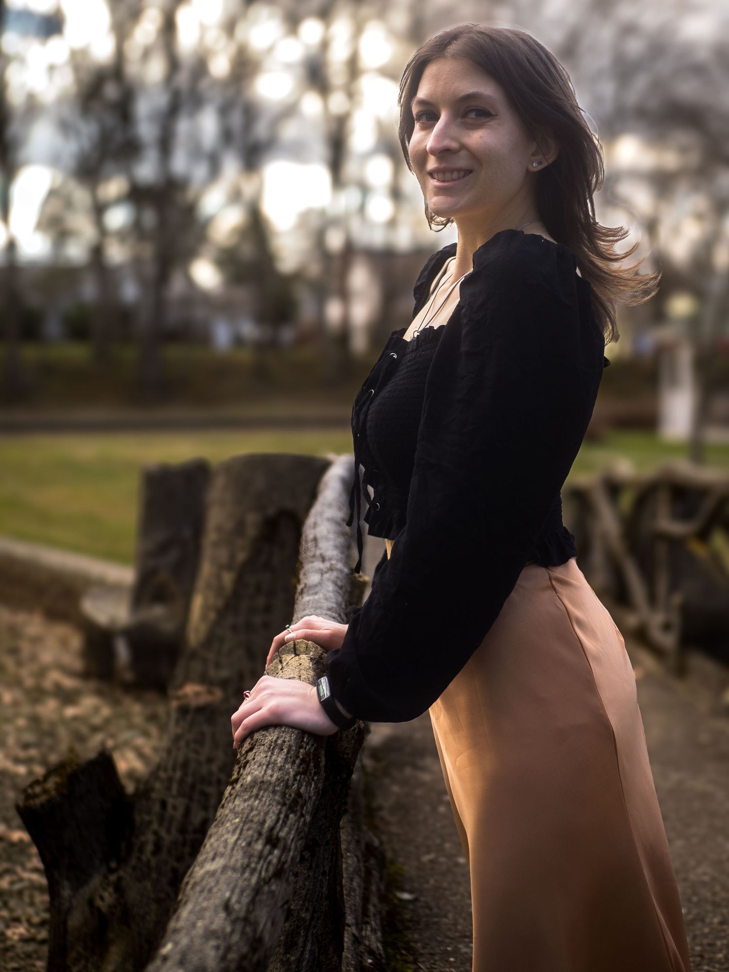 A person in a black sweater and beige skirt poses against a rustic wooden fence in an autumn garden setting.