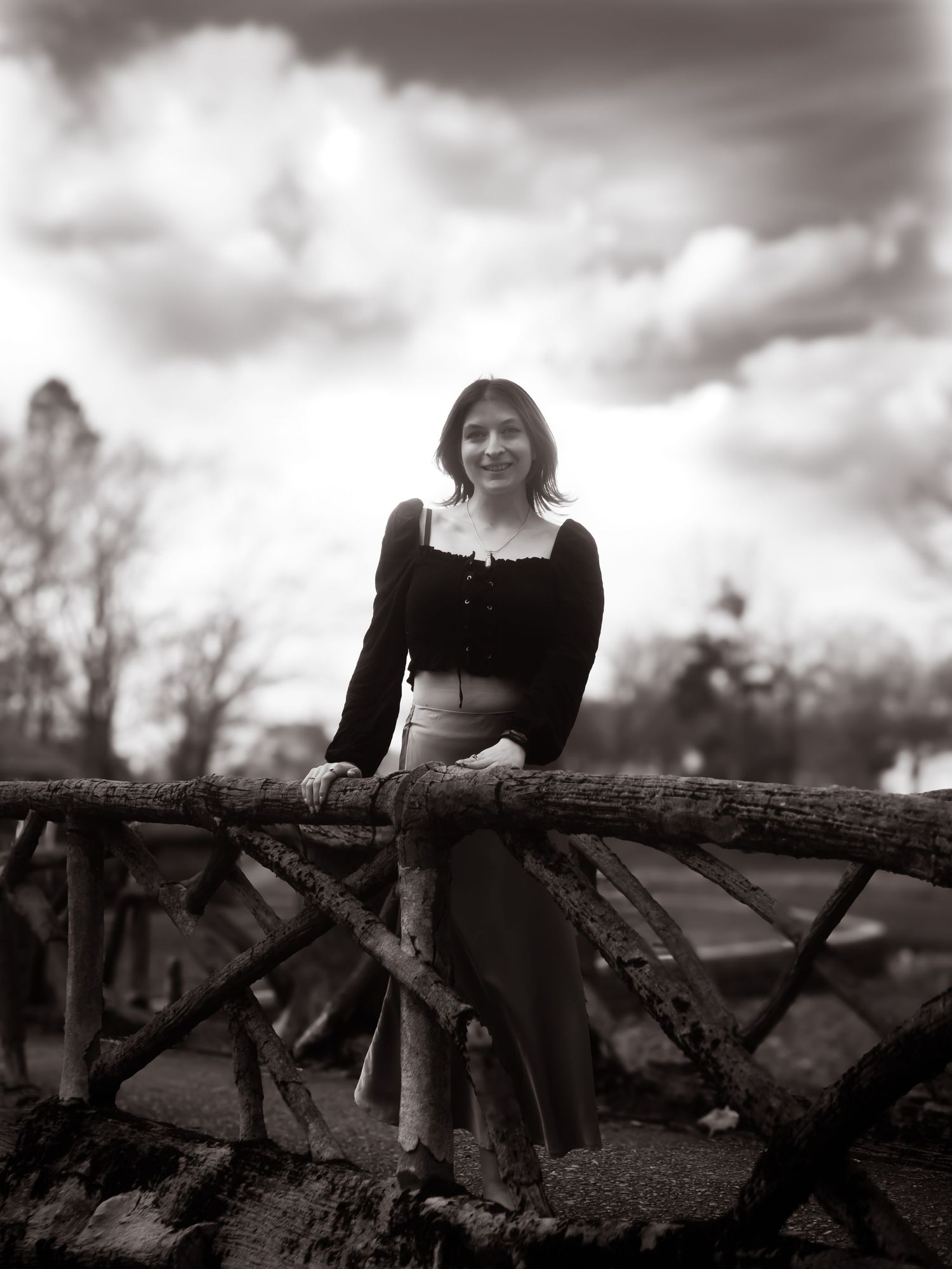 Black and white vintage style photo of someone sitting on a rustic wooden bridge under dramatic cloudy skies.