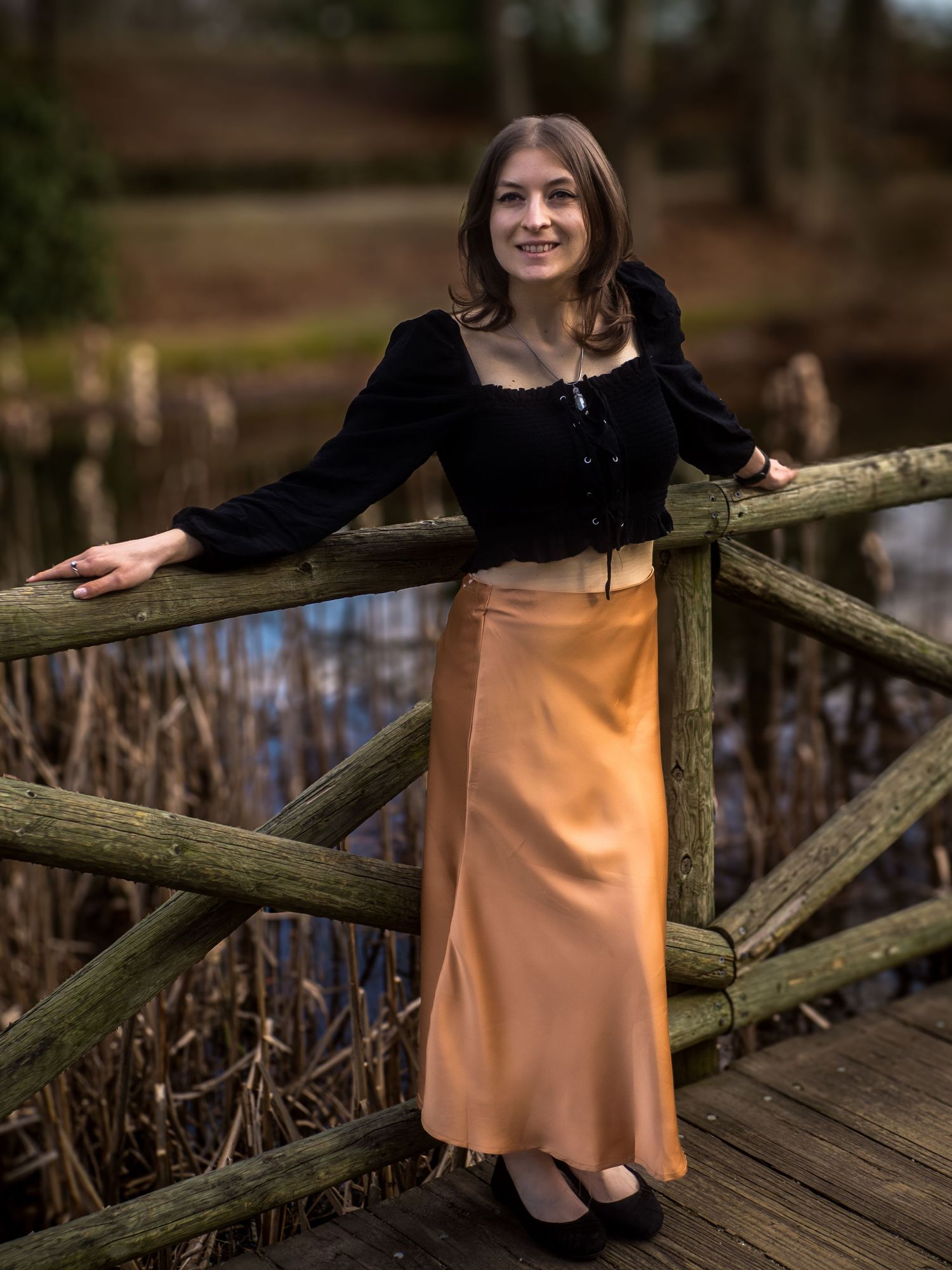 Woman in black velvet top and peach satin skirt poses on wooden bridge in outdoor nature setting.