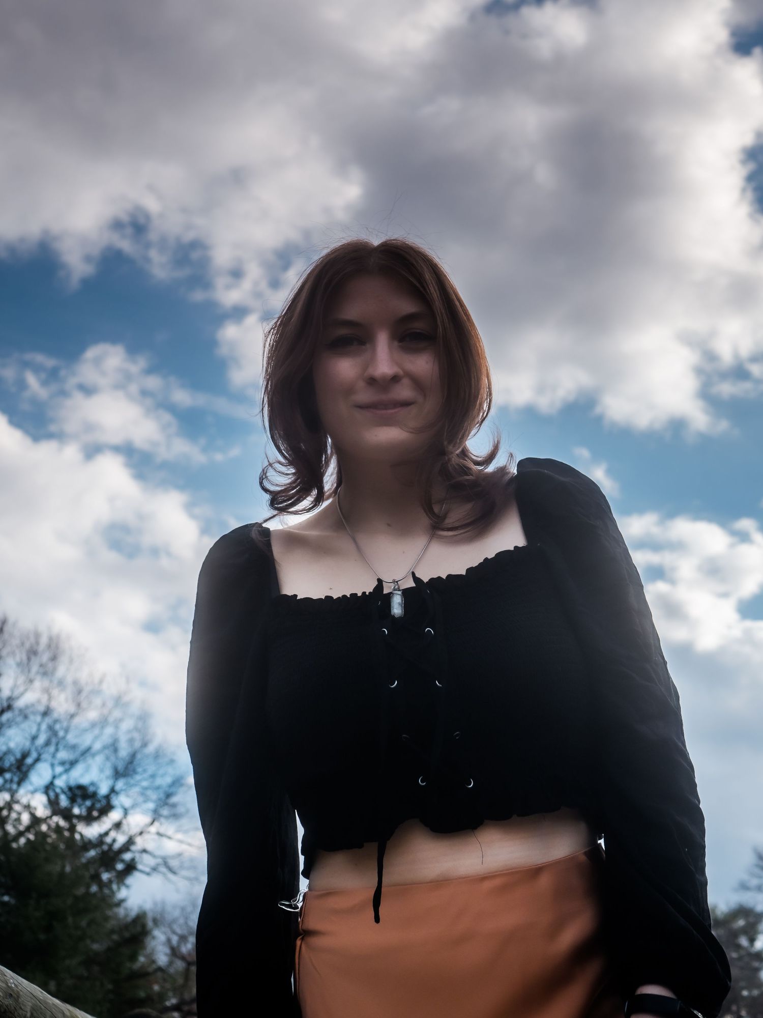 A person in a black crop top poses outdoors under a dramatic cloudy sky.